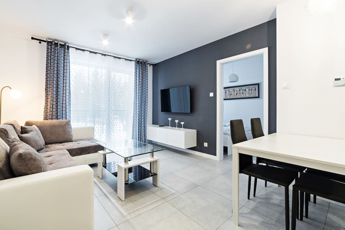 A modern living room with a sofa, a glass coffee table, a dining table with chairs, a TV mounted on the wall, and a door leading to another room.