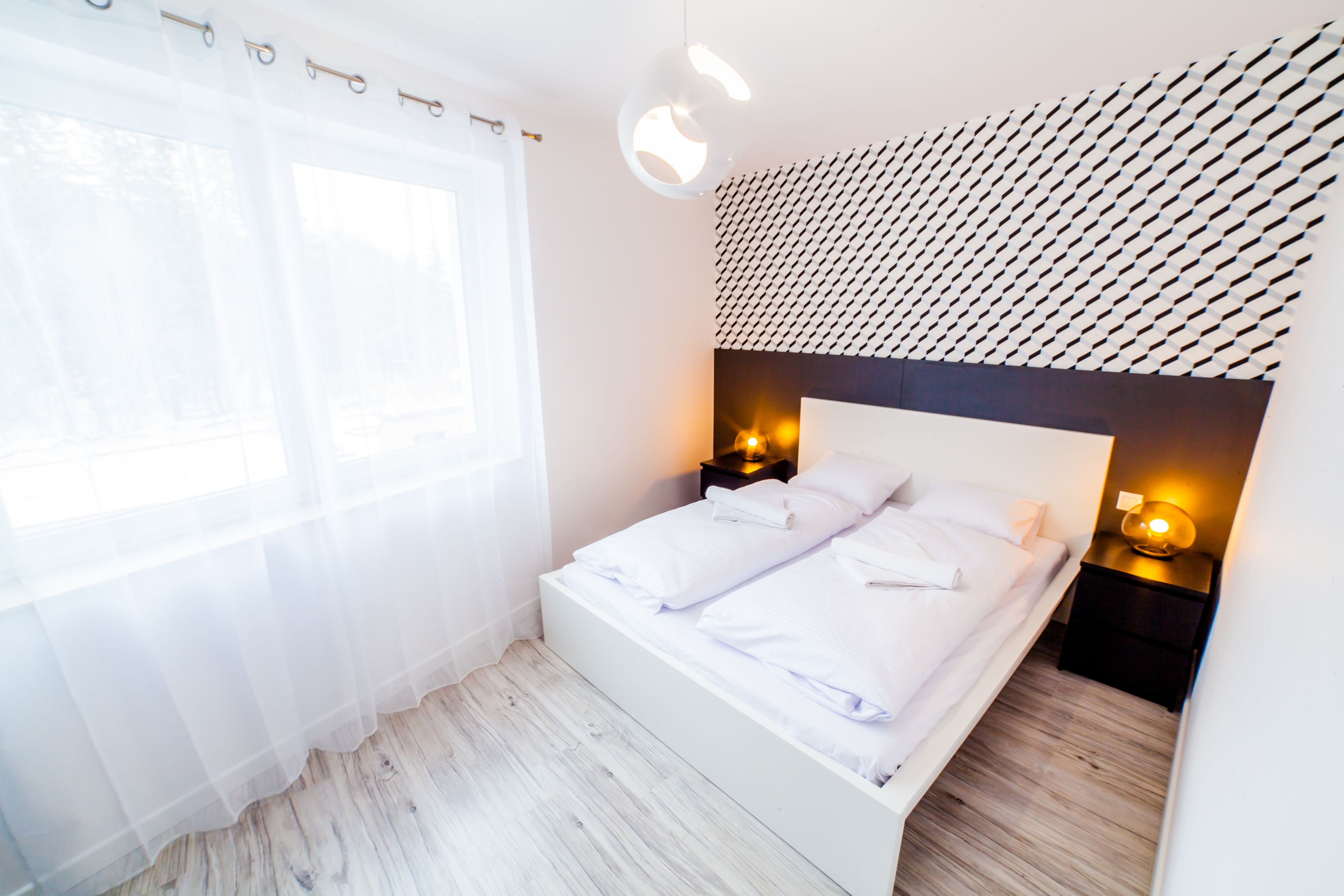 Quarto moderno com cama branca, duas mesas de cabeceira com abajures, cortinas leves e parede estampada.