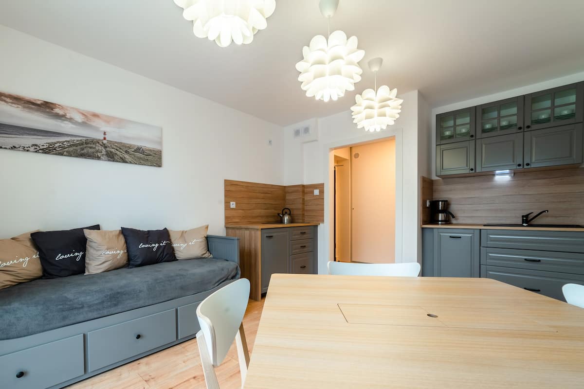 A modern living room with a grey sofa, wooden dining table, and a kitchen area with grey cabinets and a coffee machine.