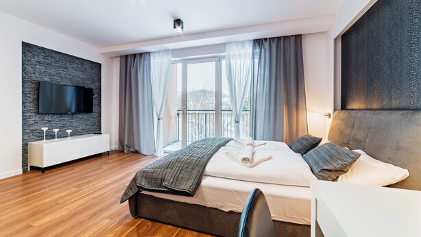Una habitación de hotel moderna con una cama grande, un televisor de pantalla plana montado en una pared de piedra, un balcón con vistas y una mesita de noche blanca.