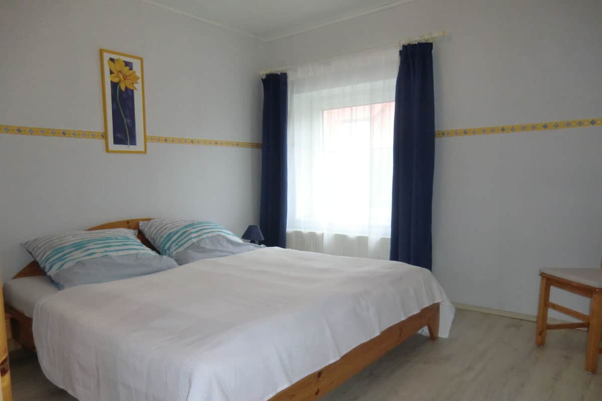 Un dormitorio con cama de madera, dos almohadas, una mesita de noche, una lámpara y una ventana con cortinas.