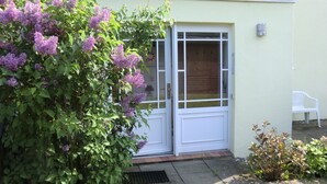 A house with a white door and window, a small garden with purple flowers, and a white plastic chair outside. - Böltser Hus (Neukirchen)