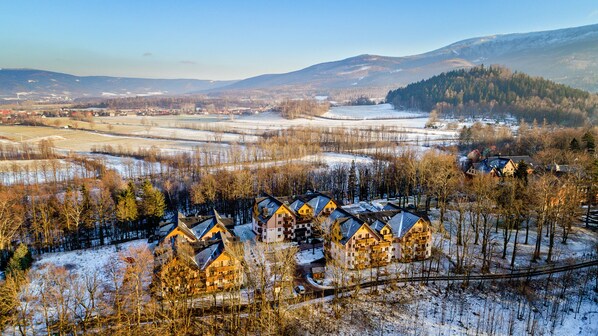 A cluster of traditional wooden houses with sloped roofs, surrounded by snow-covered fields and trees, set against a backdrop of mountains. - Apartamenty Sun&Snow Lesny Dom (Karpacz)