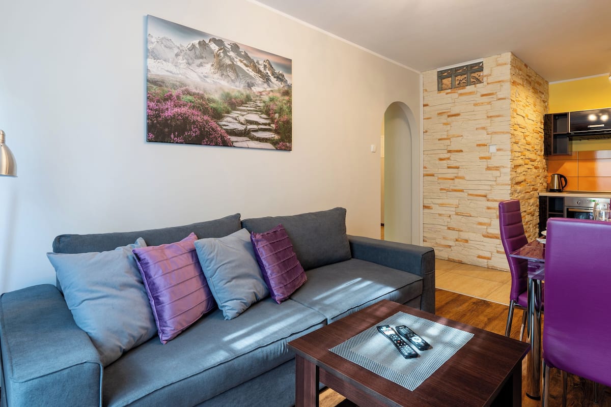 Una sala de estar moderna con un sofá gris, cojines morados y una mesa de centro con controles remotos. Un cuadro de un paisaje montañoso cuelga en la pared.
