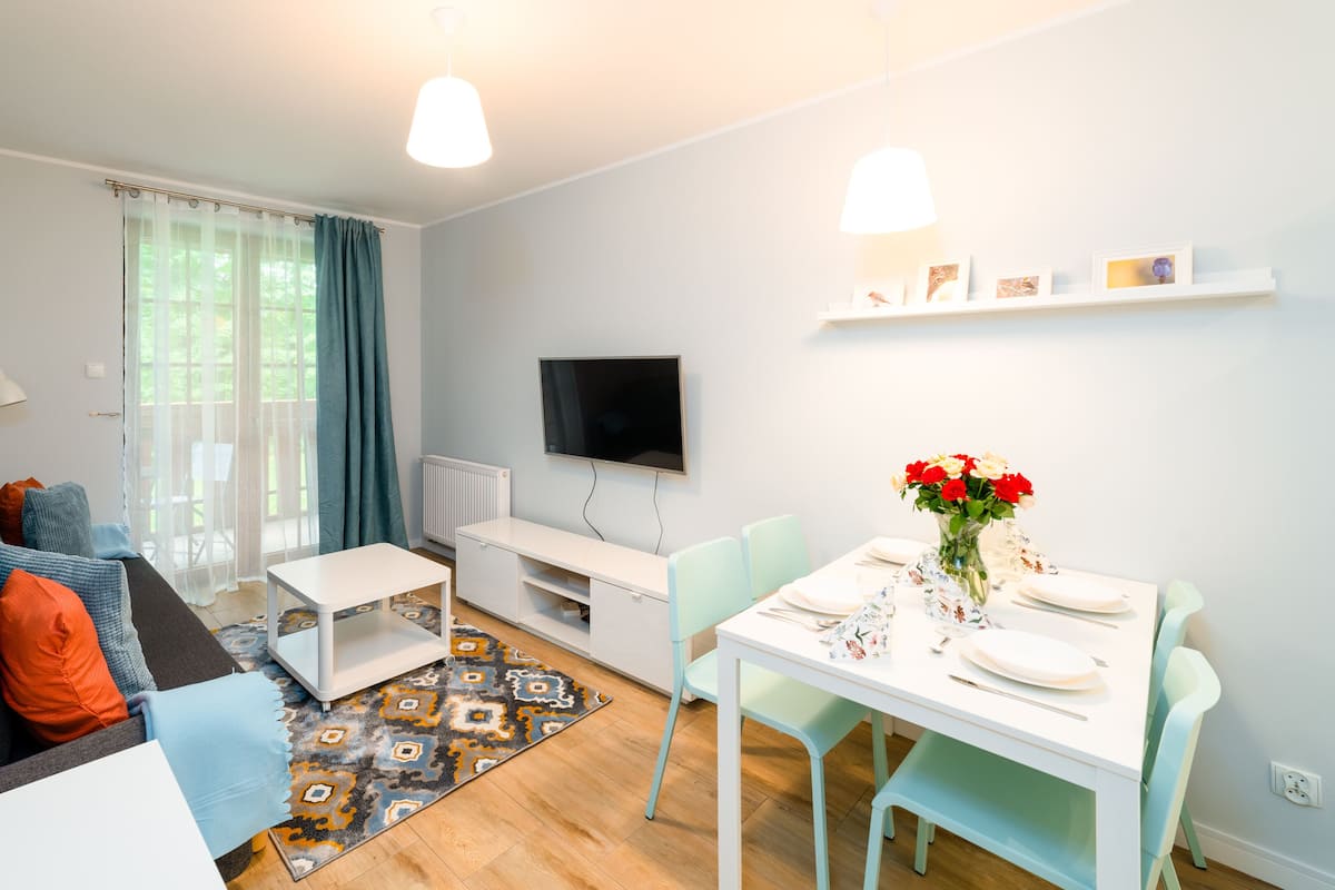 Una sala moderna con un sofá, una mesa de comedor preparada para dos personas, un televisor en la pared y una ventana con cortinas.