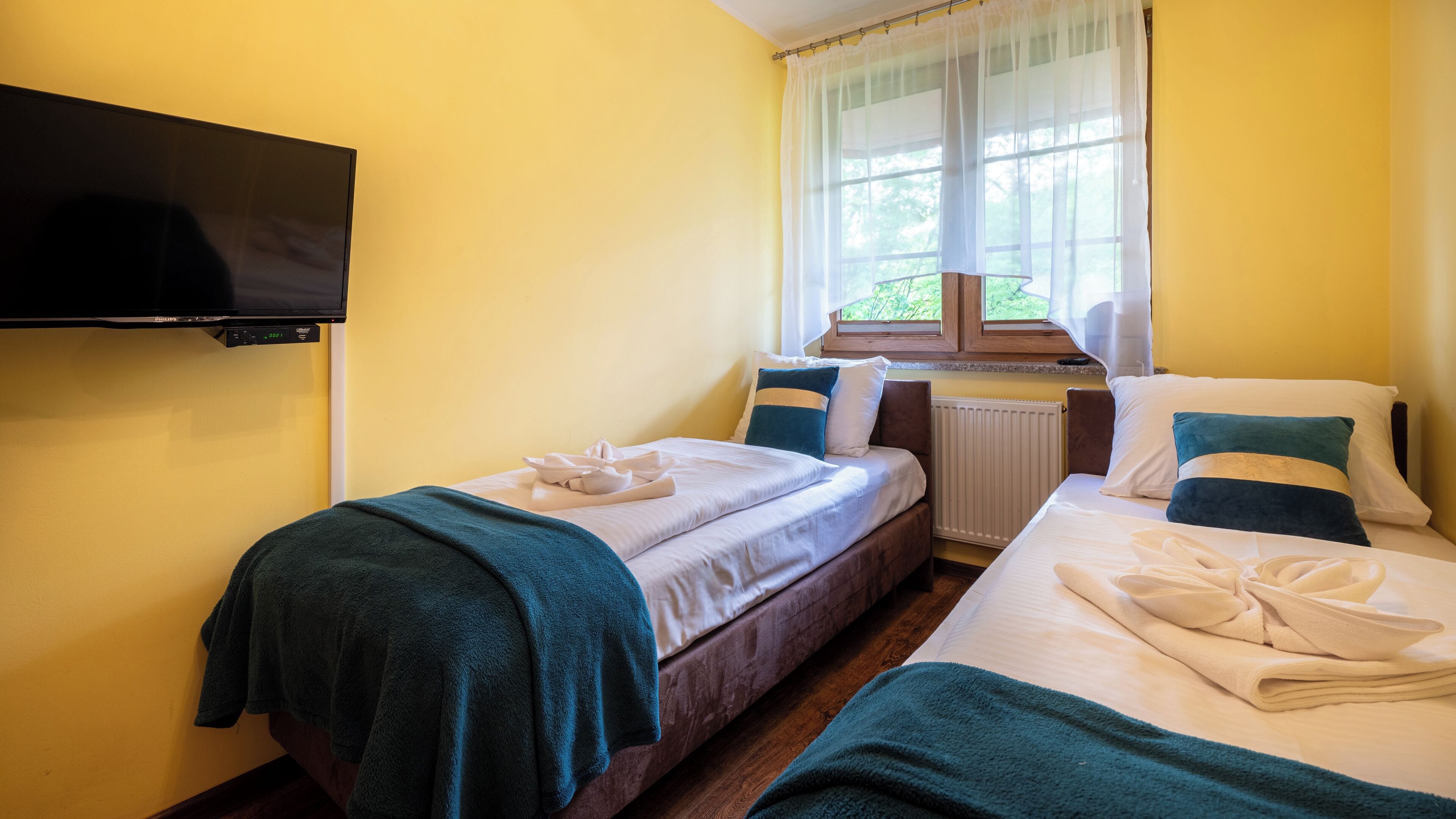A hotel room with two beds, a TV mounted on the wall, a window with curtains, and a radiator.