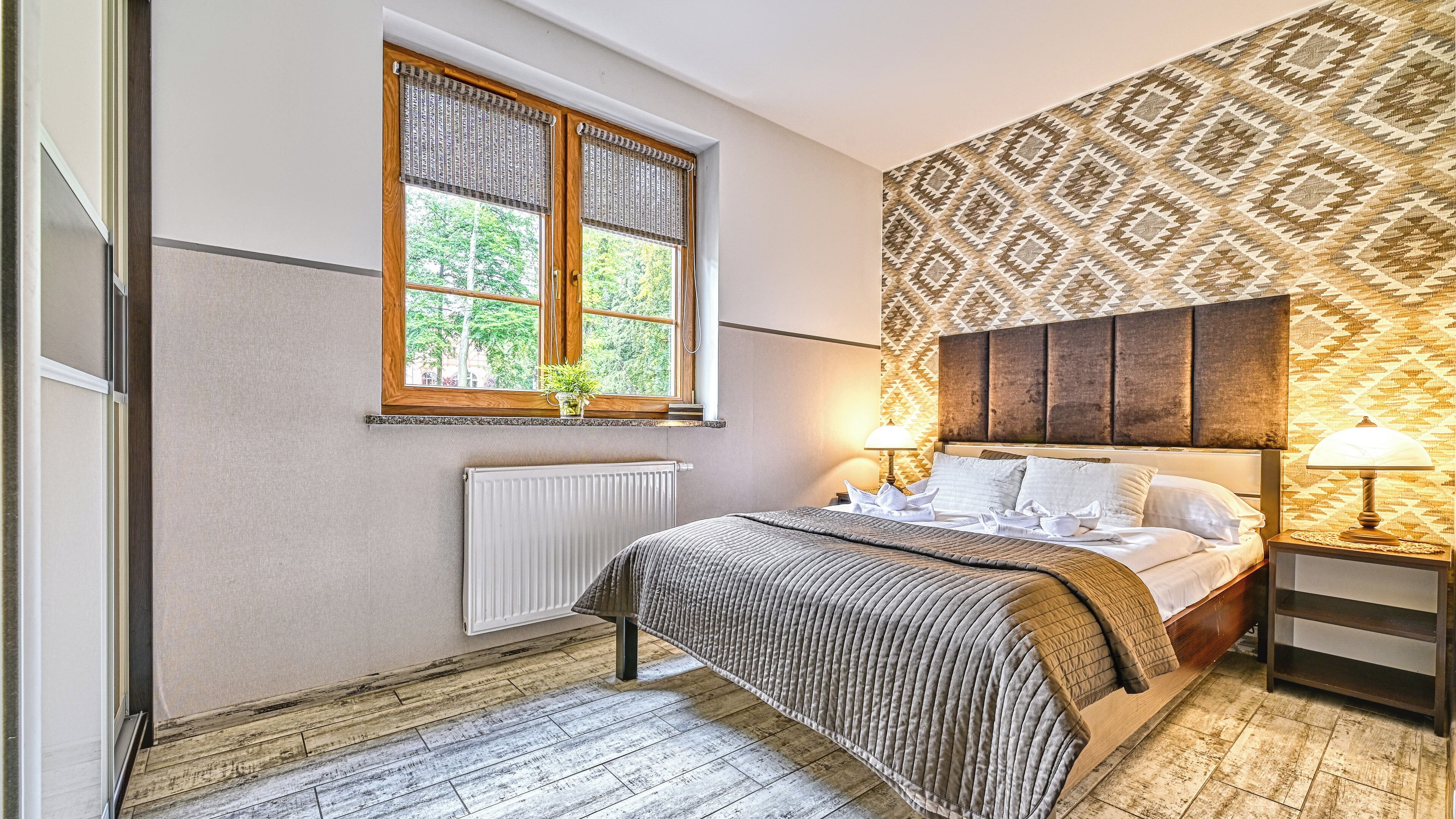Ein Schlafzimmer mit einem Bett, zwei Nachttischlampen, einem Fenster mit Blick auf Bäume, einer Heizung und ornamentalem Wandpapier.