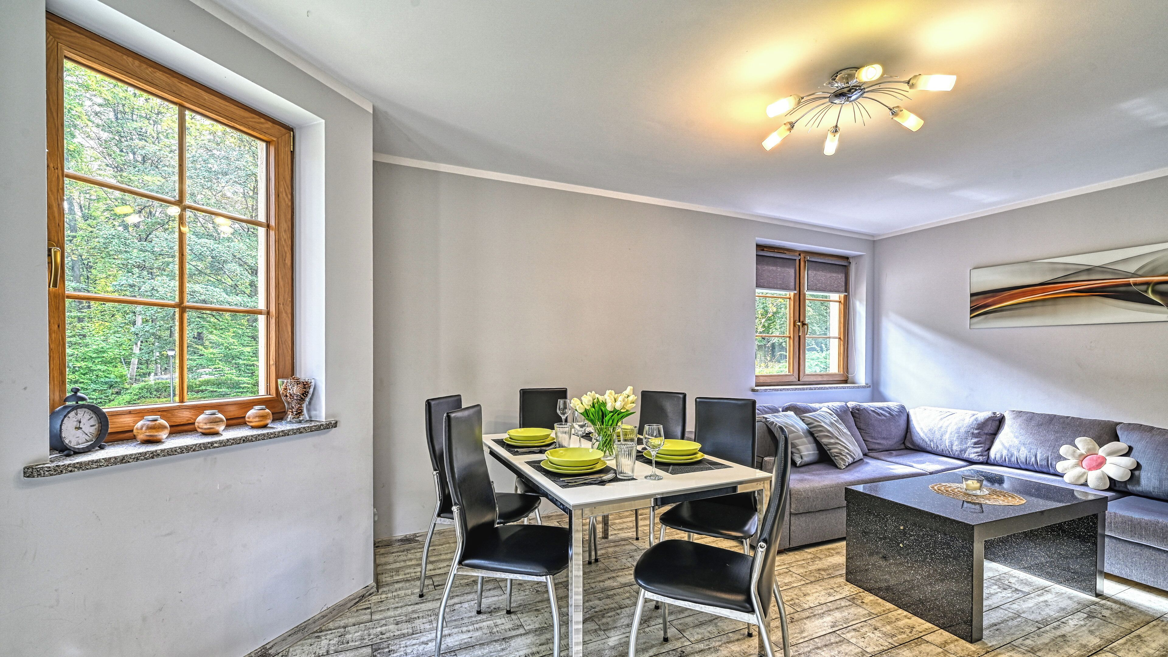 A dining area with a table set for four, chairs, a sofa, and a coffee table. There are two windows providing natural light, and a ceiling light fixture.