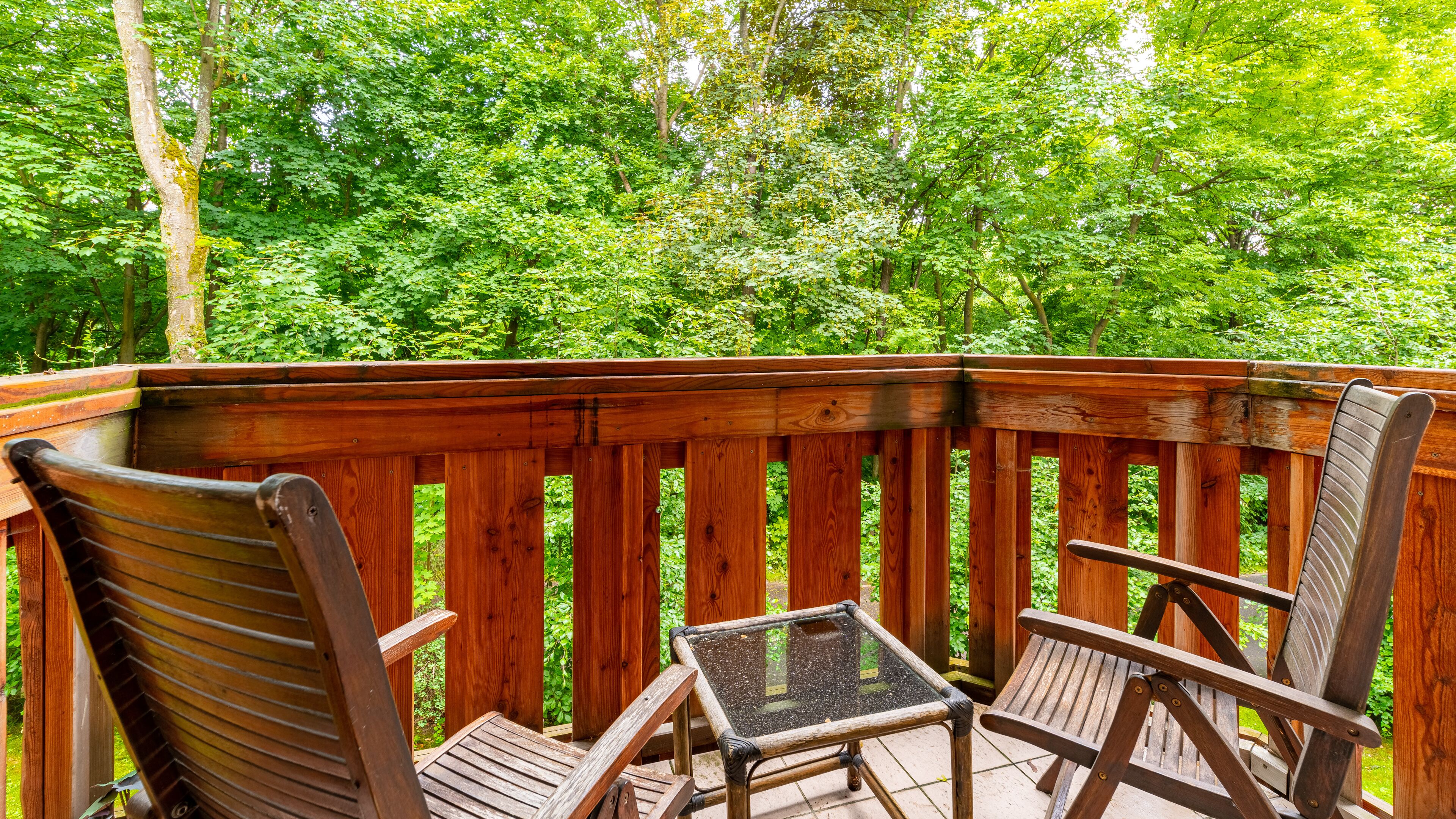 A wooden deck with two wooden chairs and a small table, surrounded by lush green trees.