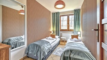 A room with two beds, a mirror, a window with curtains, a radiator, and a hanging light fixture.