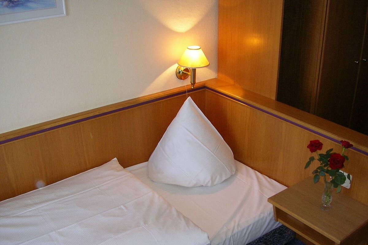 A neatly made bed with white linens, a single white pillow, and a small bedside table with a vase of red roses.