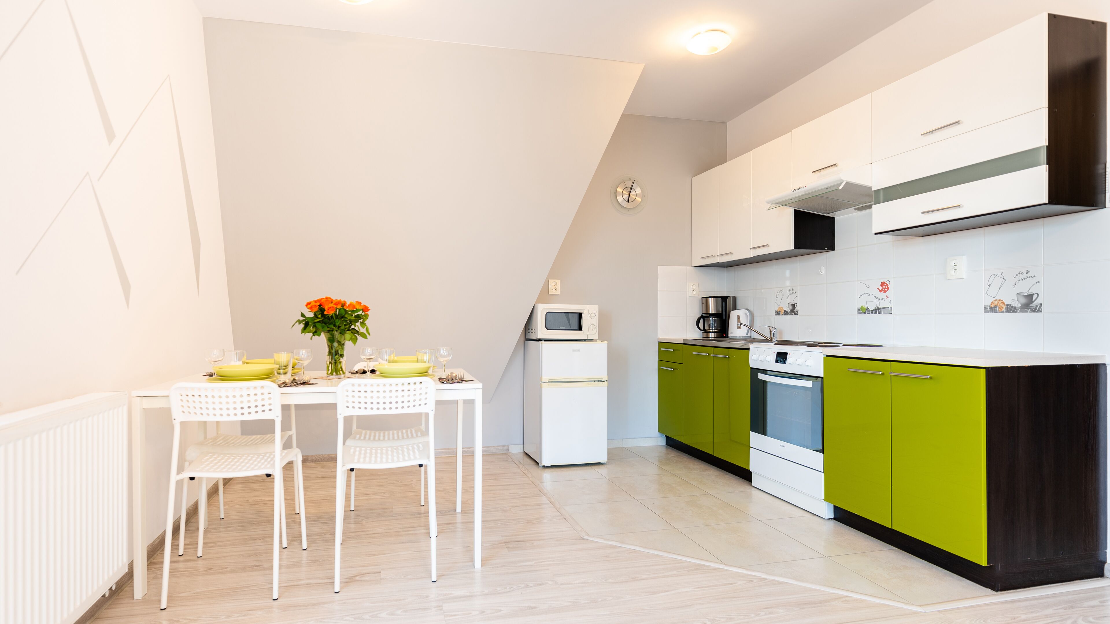 Une cuisine moderne avec des meubles blancs et verts, un réfrigérateur blanc, un four à micro-ondes, une machine à café et une cuisinière. On y trouve une table à manger avec des chaises et un vase de fleurs.