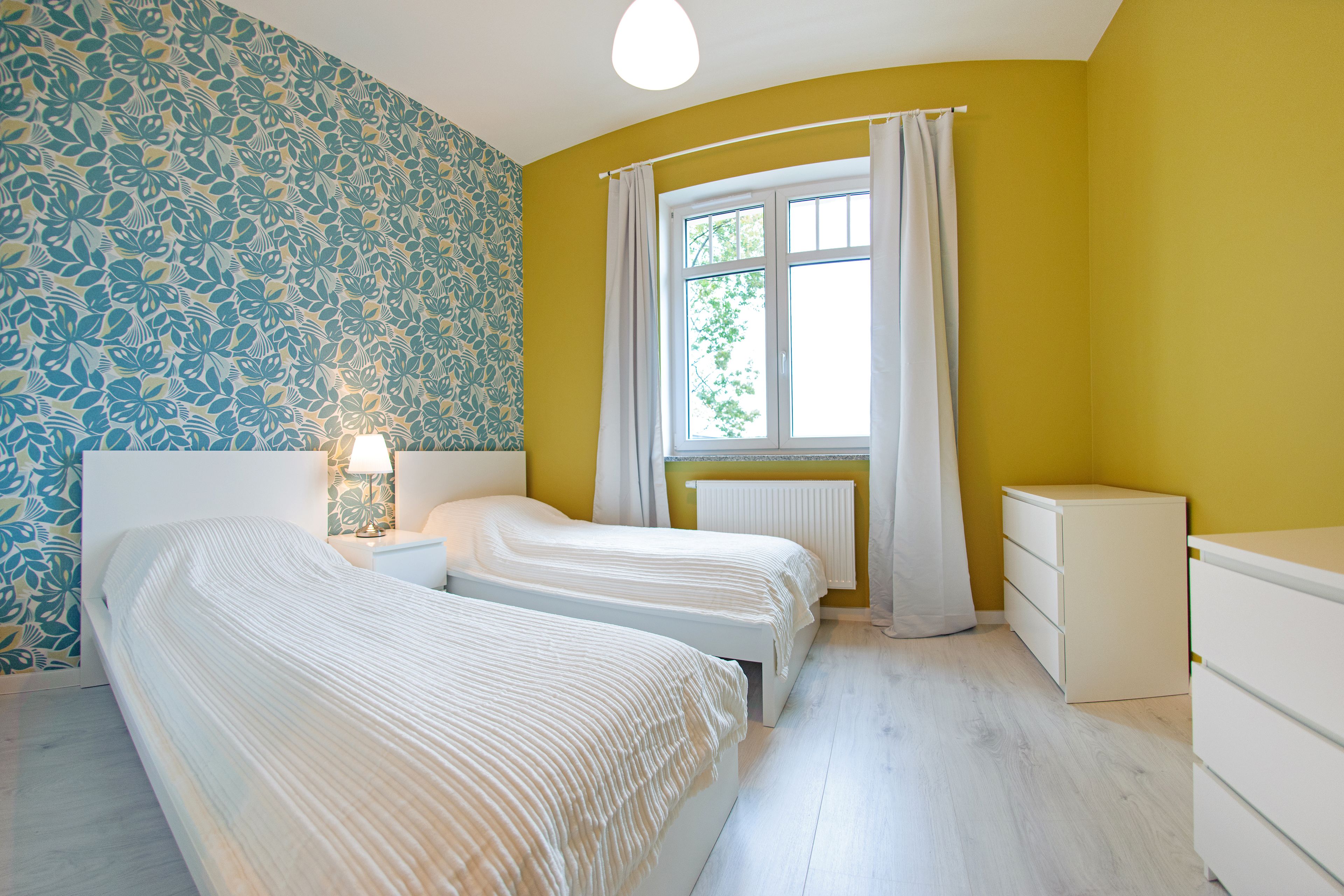 Quarto com duas camas, uma janela com cortinas, um radiador e uma parede estampada.