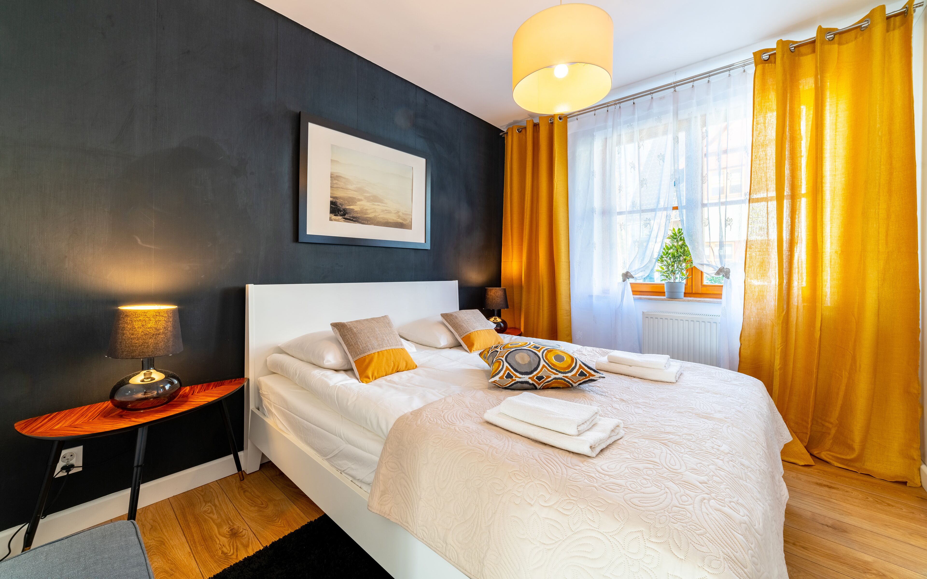 A neatly made bed with white linens and decorative pillows, a bedside lamp, a framed artwork on the wall, and a window with yellow curtains.
