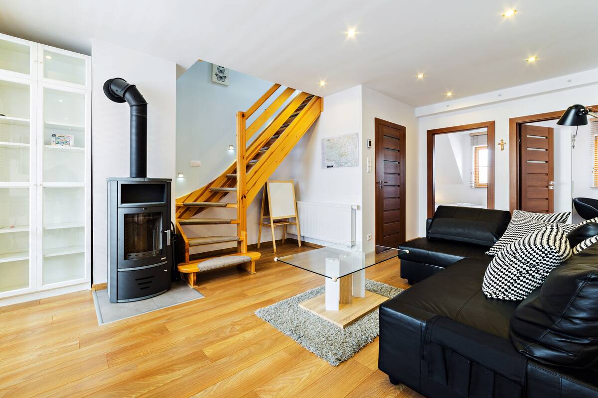 Ein modernes Wohnzimmer mit einem schwarzen Kaminofen, einem Glastisch, einem schwarzen Ledersofa, Holzböden und einer Holztreppe.