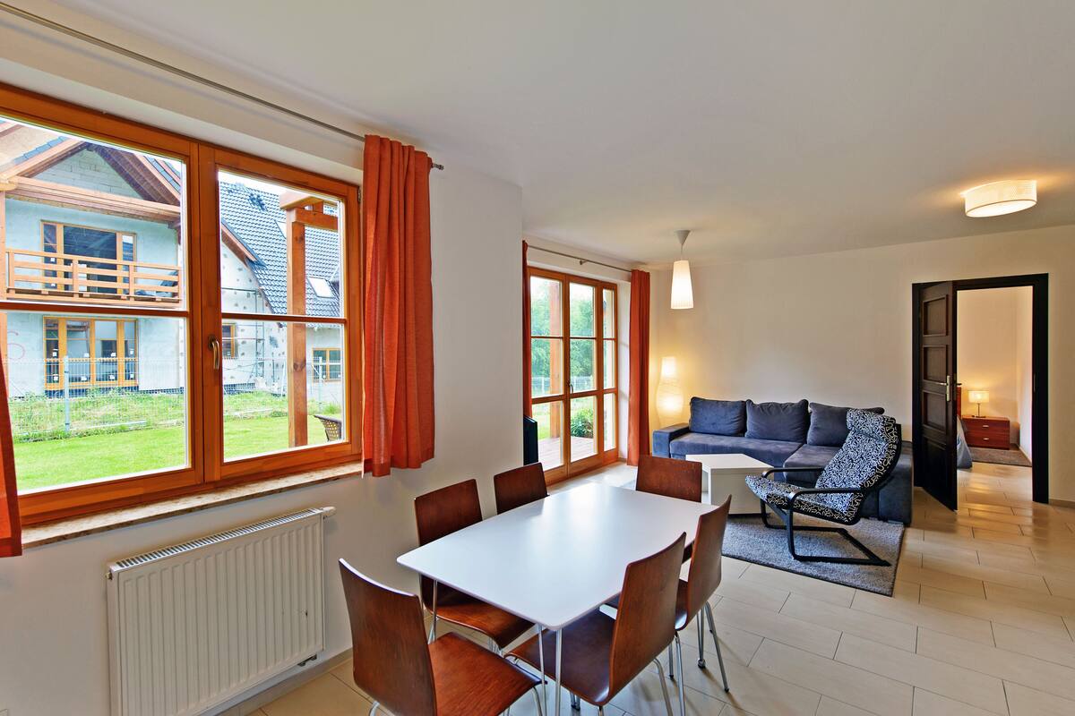 Ein Essbereich mit einem weißen Tisch und braunen Stühlen, einer Couch mit gemusterten Kissen und großen Fenstern mit Vorhängen.