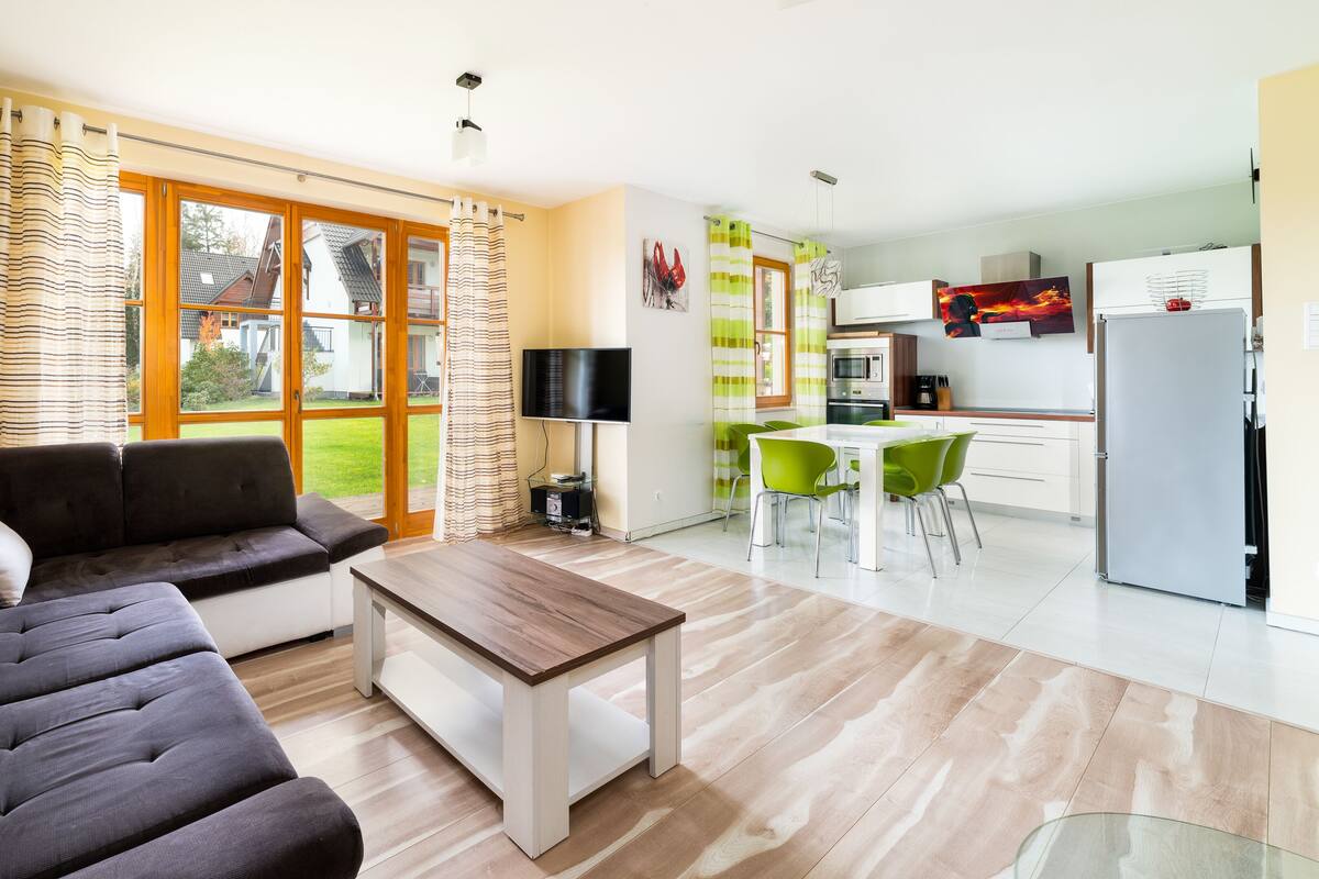 Ein modernes Wohnzimmer mit einer Couch, einem Couchtisch und einem Fernseher. Es gibt eine Küchenzeile mit einem Esstisch und Stühlen.