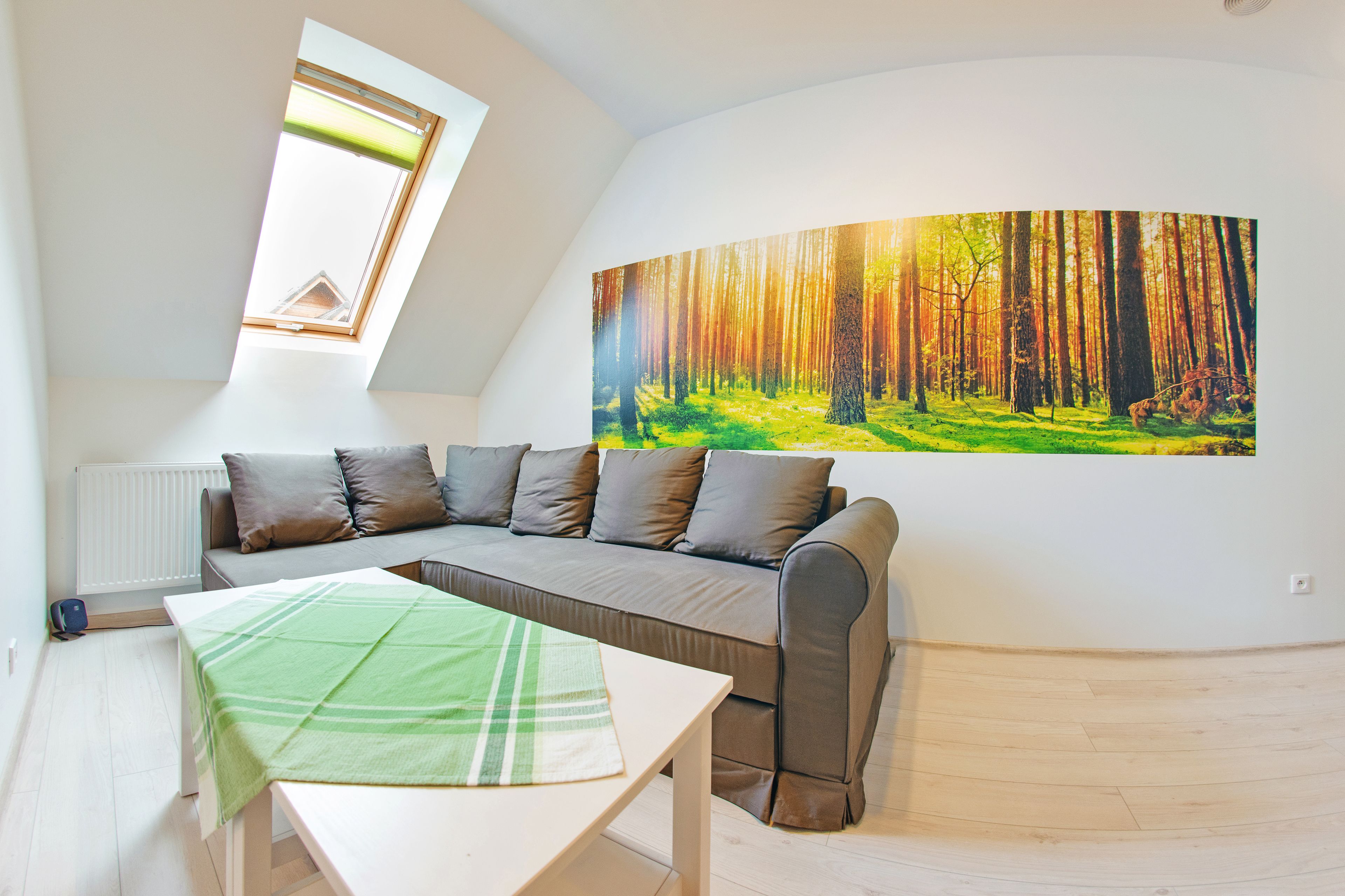 A modern living room with a sloped ceiling, a large sofa, a small table, and a forest mural on the wall.