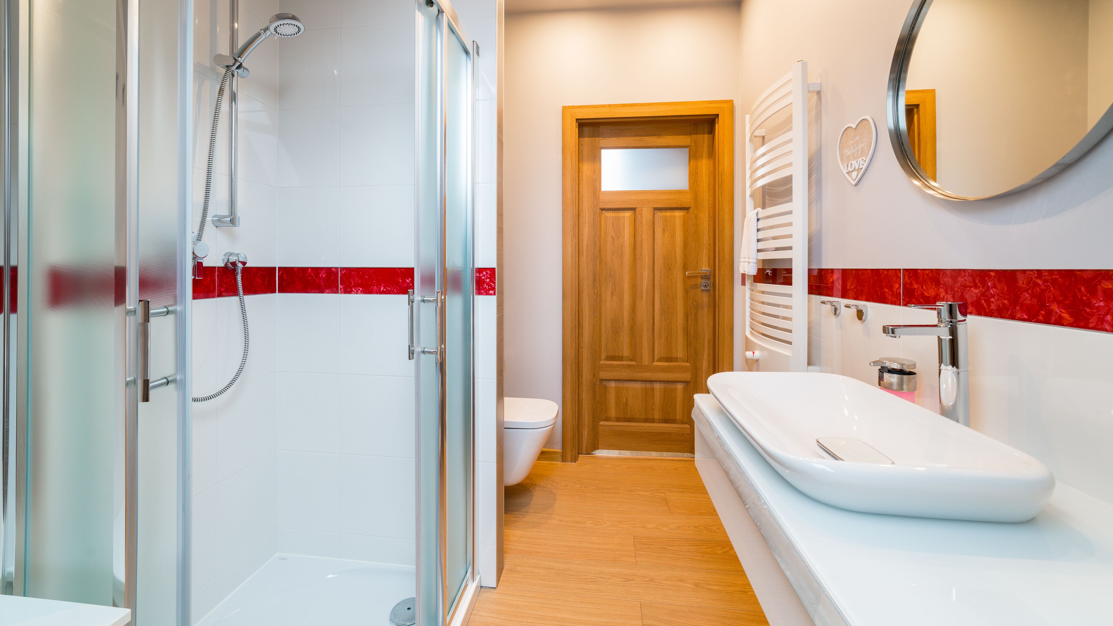 A modern bathroom with a glass shower enclosure, a wooden door, a white sink, and a red-tiled shower area.