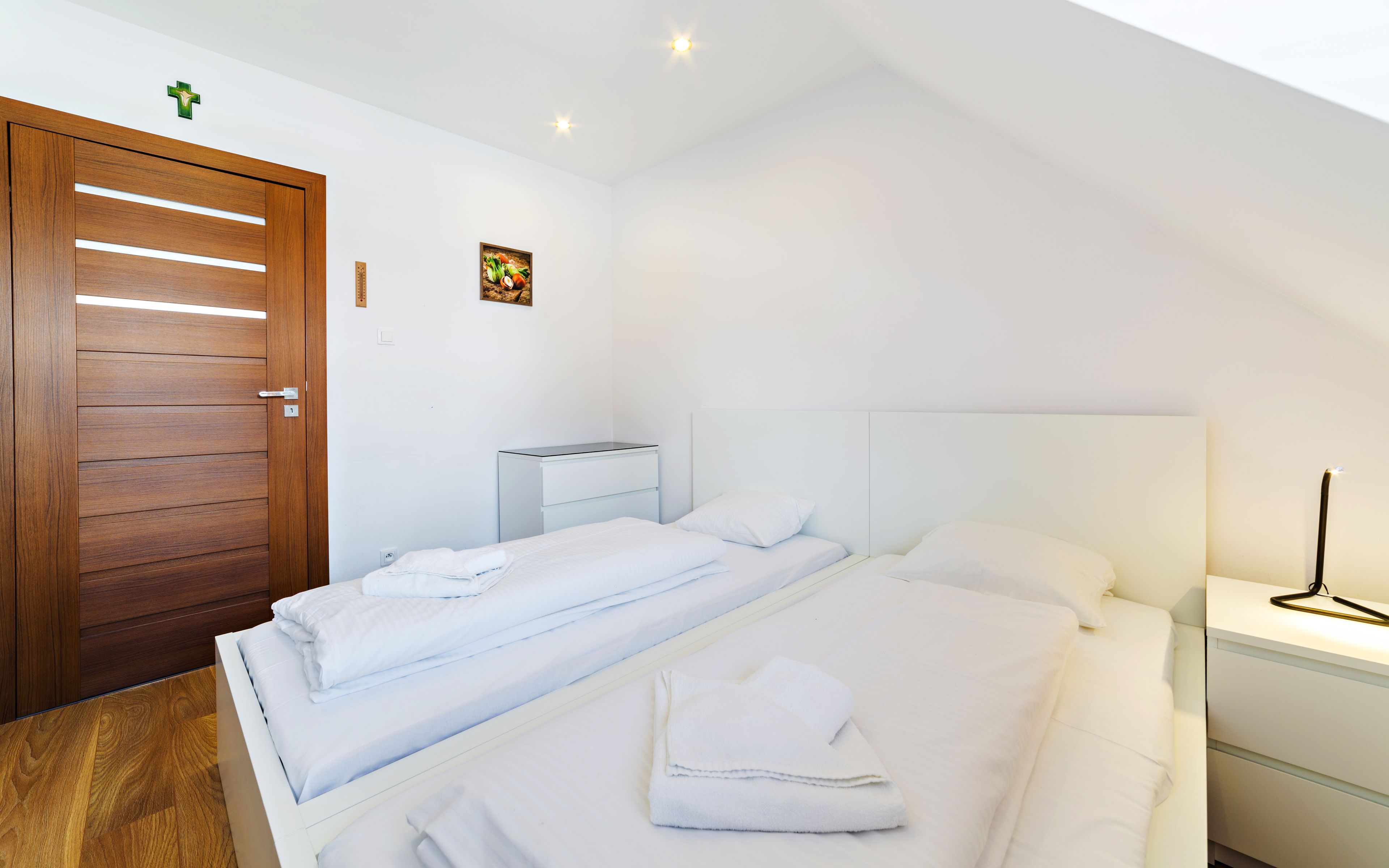 A bedroom with two beds, a wooden door, a small bedside table, and a wall-mounted light fixture.