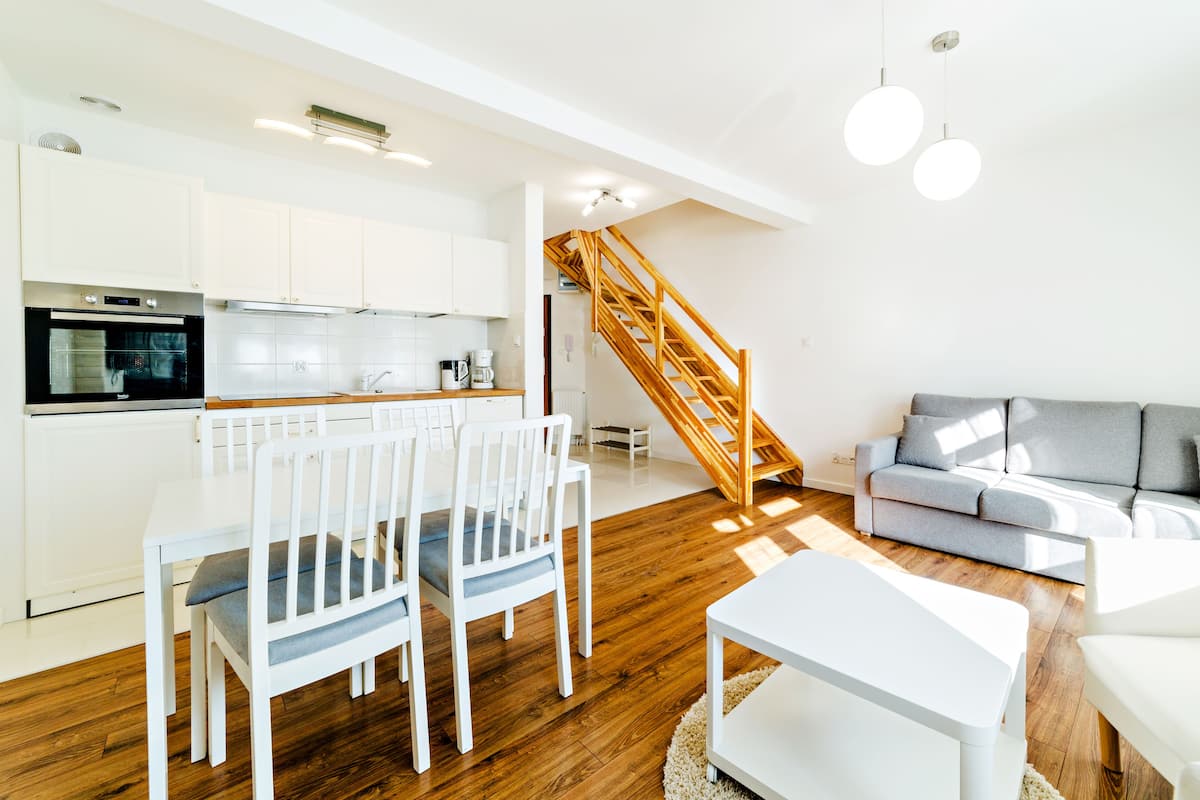 Een moderne keuken met witte kasten, een ingebouwde oven, en een eetgedeelte met witte stoelen. Een trap leidt naar een woonruimte met een grijze bank en een witte salontafel.