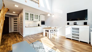 A modern kitchen with white cabinets, a dining area with a wooden table and chairs, a large flat-screen TV mounted on the wall, and a staircase leading to an upper level.
