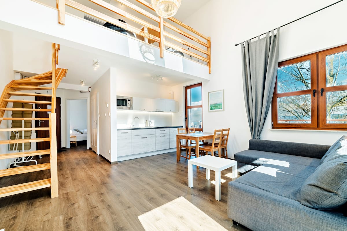 Een ruim loft appartement met een houten trap, een keuken met witte kasten, een eethoek en een groot raam met gordijnen.