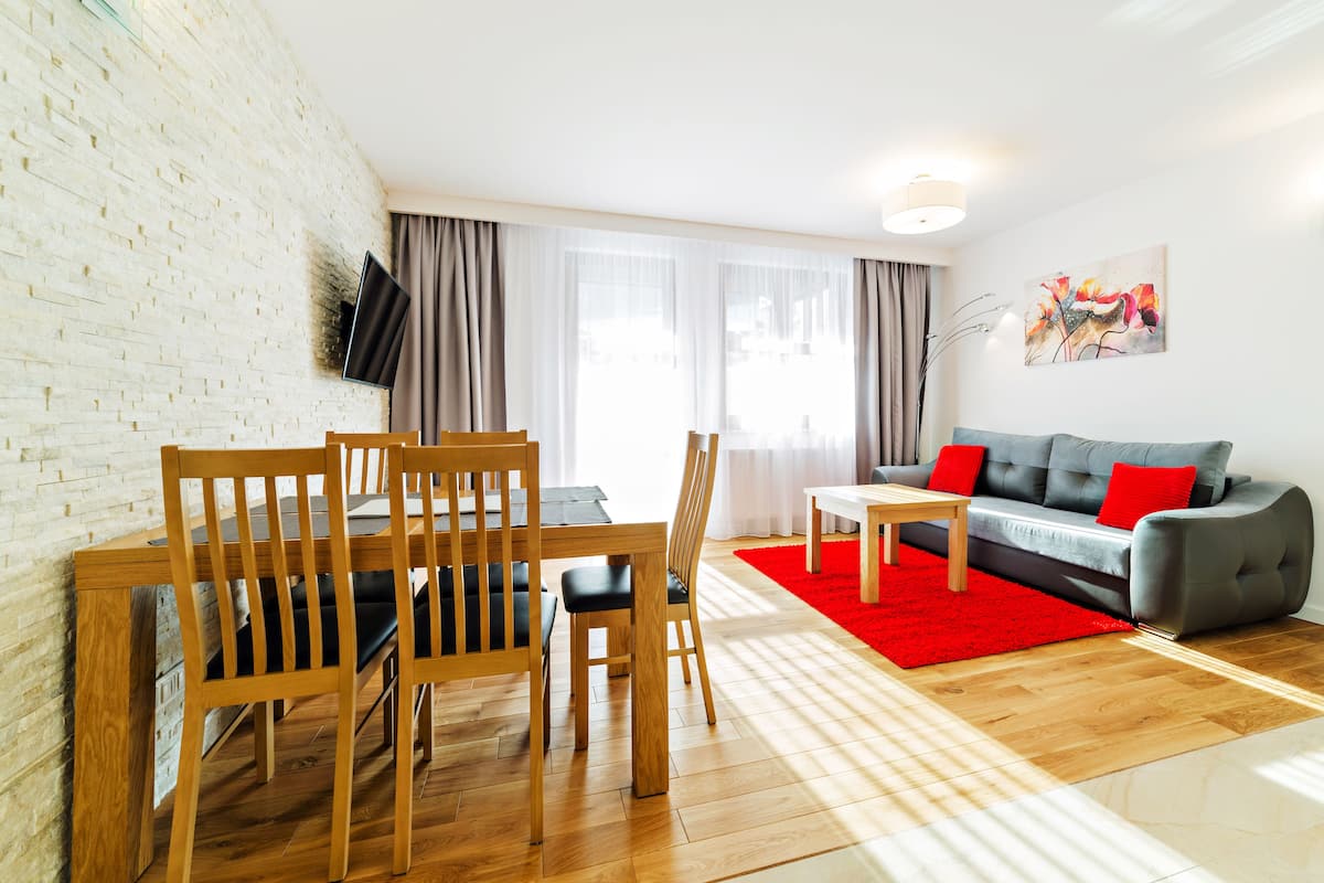 Een woonkamer met een eethoek, een grijze bank met rode kussens, een rood tapijt en een bakstenen muur.