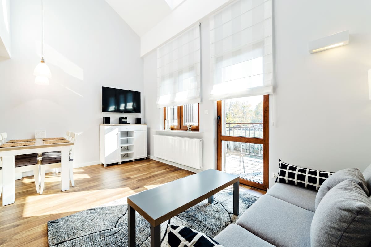 Een moderne woonkamer met een houten vloer, een grijze bank, een eettafel met stoelen, een televisie aan de muur en grote ramen met luxaflex.