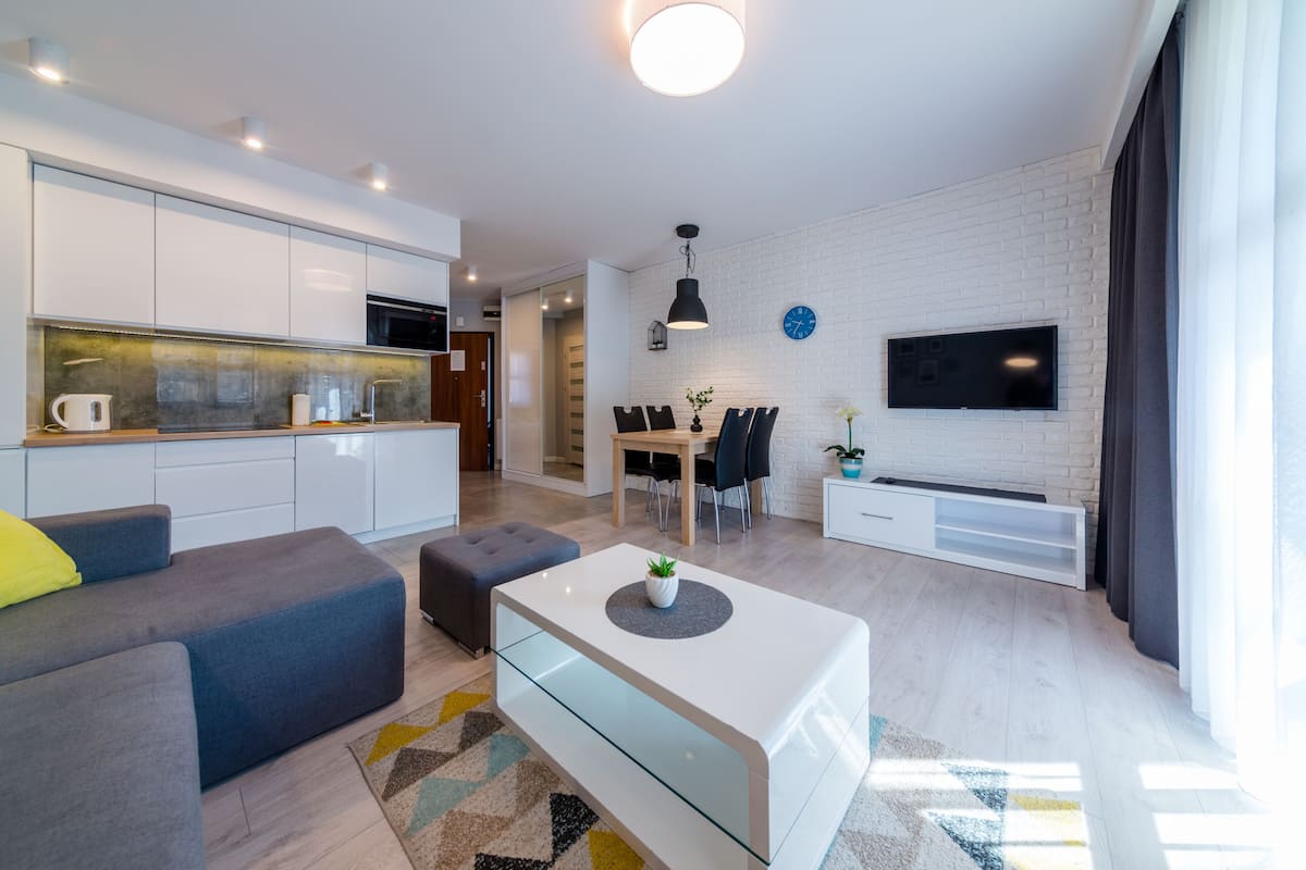 Een moderne woonkamer met een bank, een salontafel en een televisie. Er is een keukenhoek met kastjes en een magnetron.