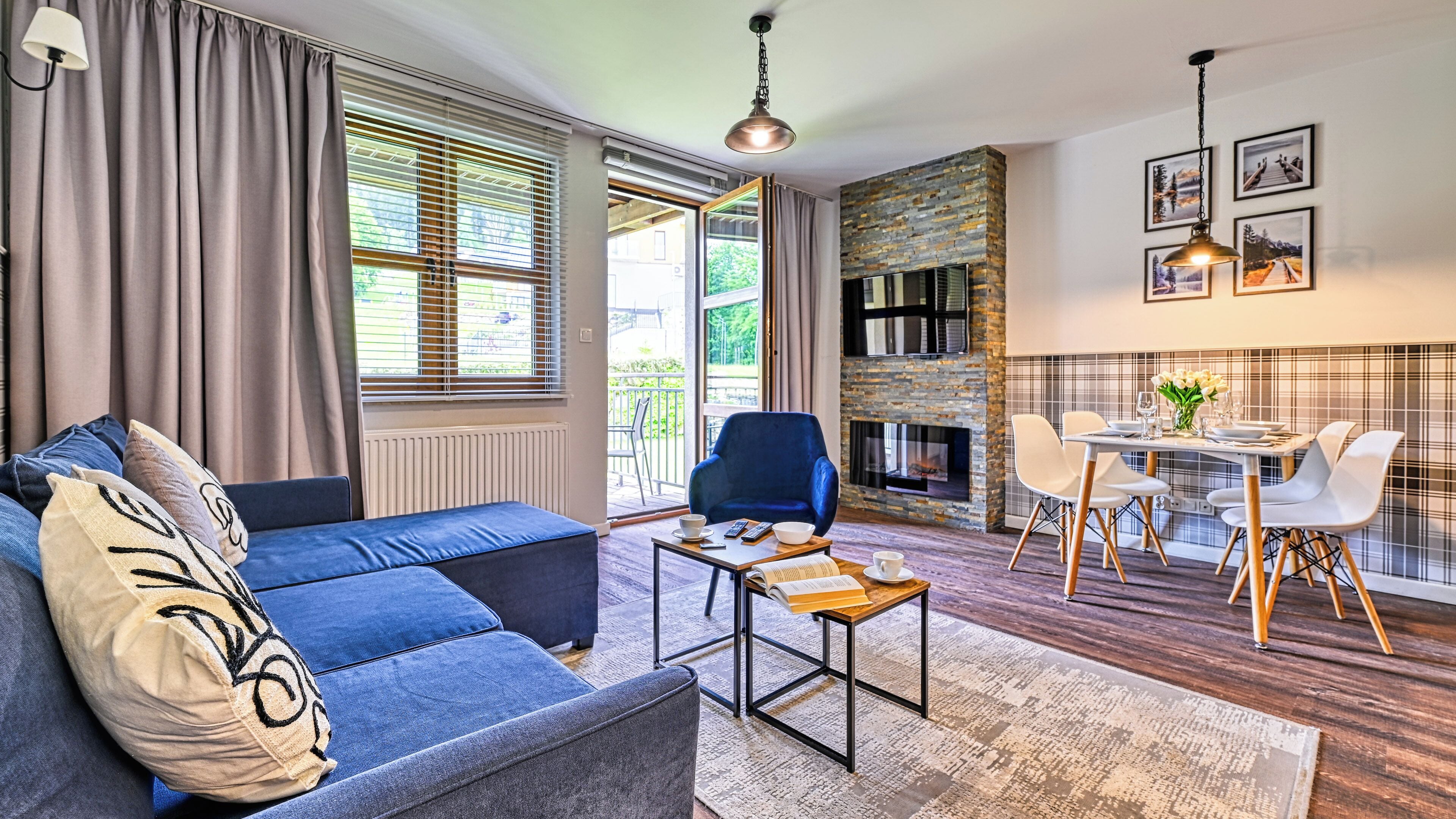 Un salon moderne avec un canapé bleu, un coin repas comprenant une table et des chaises, une cheminée et de grandes fenêtres équipées de stores.