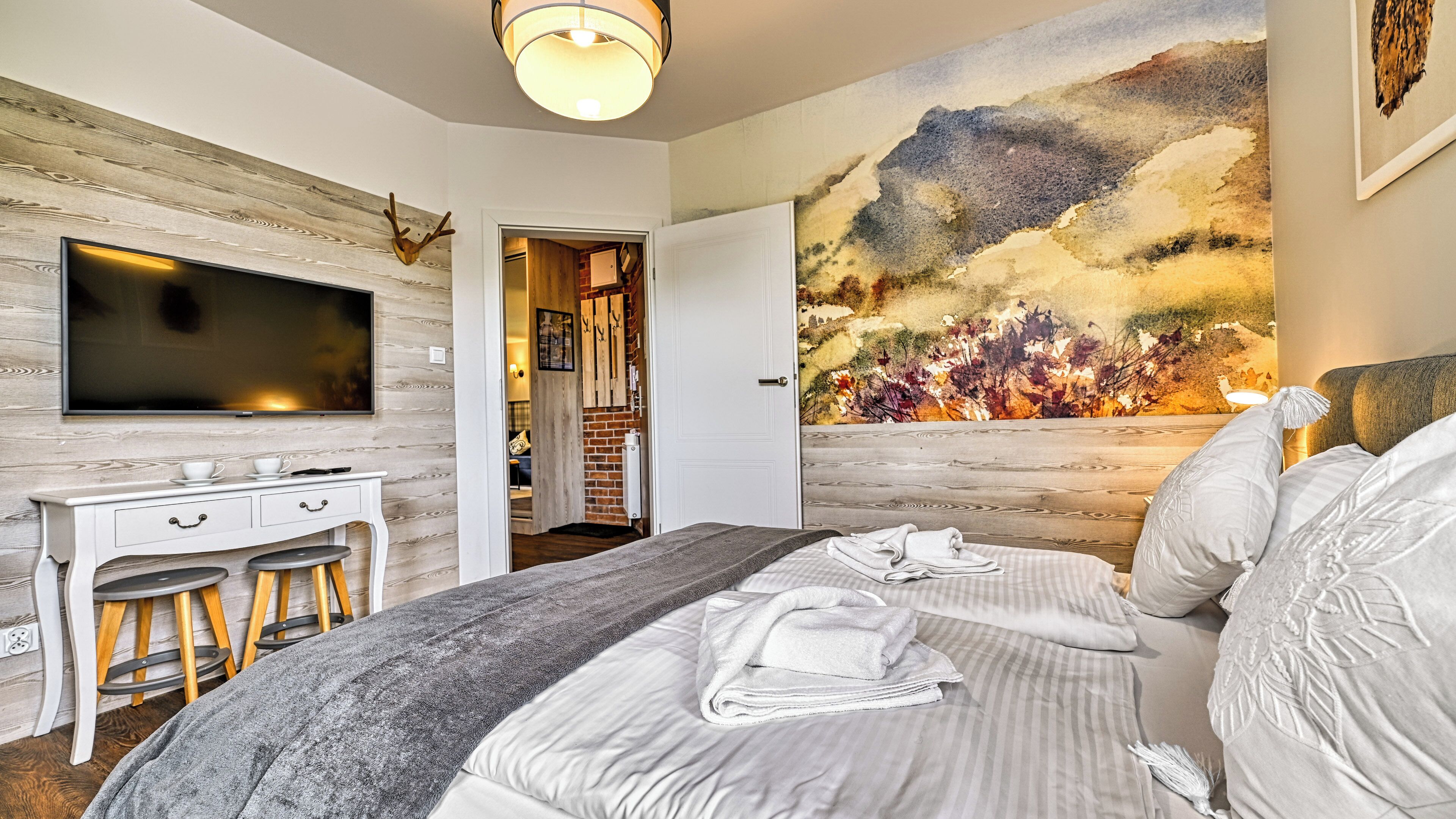 A modern bedroom with a large bed, a desk with stools, a flat-screen TV, and a large landscape painting on the wall.
