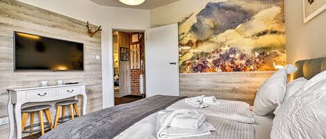 A modern bedroom with a large bed, a desk with stools, a flat-screen TV, and a large landscape painting on the wall.