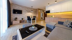 A modern living room with a flat-screen TV, a dining area with chairs, a sofa, and a kitchen with white cabinets.