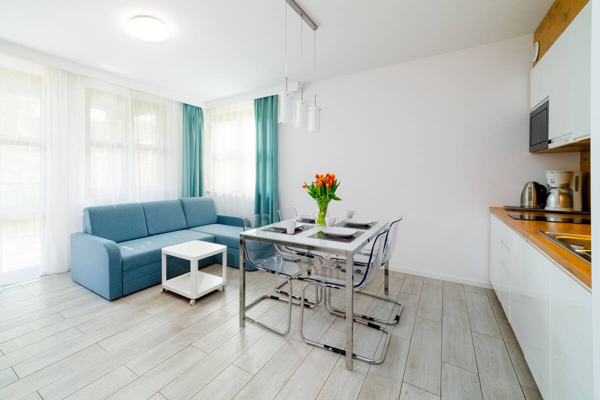 Een moderne woonkamer met een blauwe bank, een glazen eettafel en een keukenhoek met een koffiezetapparaat.