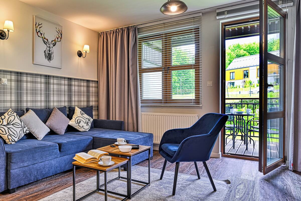 Een moderne woonkamer met een blauwe bank, een houten salontafel en een blauwe fauteuil. Er is een raam met zonwering, een radiator en uitzicht op een geel gebouw.