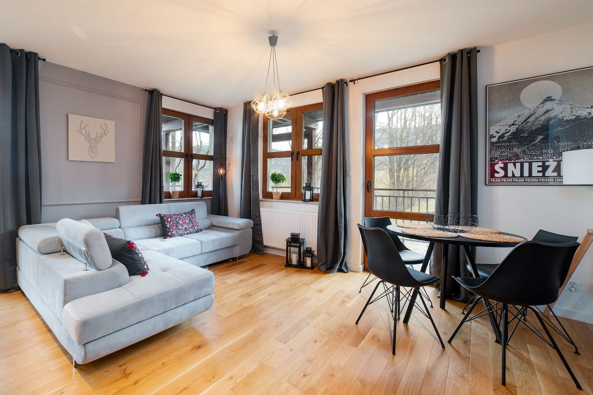 Een moderne woonkamer met een grijze bank, een eettafel met stoelen, grote ramen en uitzicht op bomen.