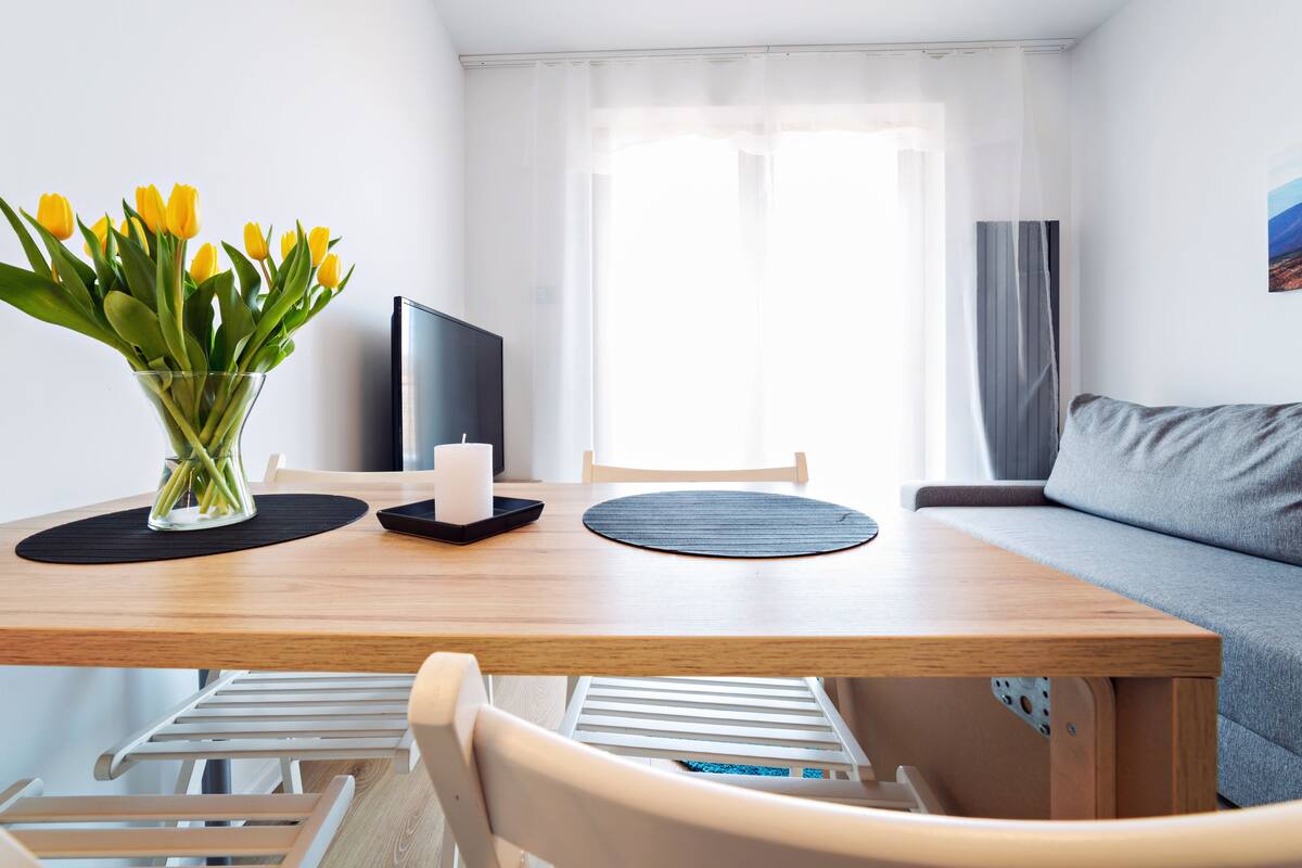 Ein modernes, gut beleuchtetes Zimmer mit einem Holzespeisetisch, einer Vase mit gelben Tulpen, einem Flachbildfernseher, einem grauen Sofa und einem Fenster mit durchsichtigen Vorhängen.