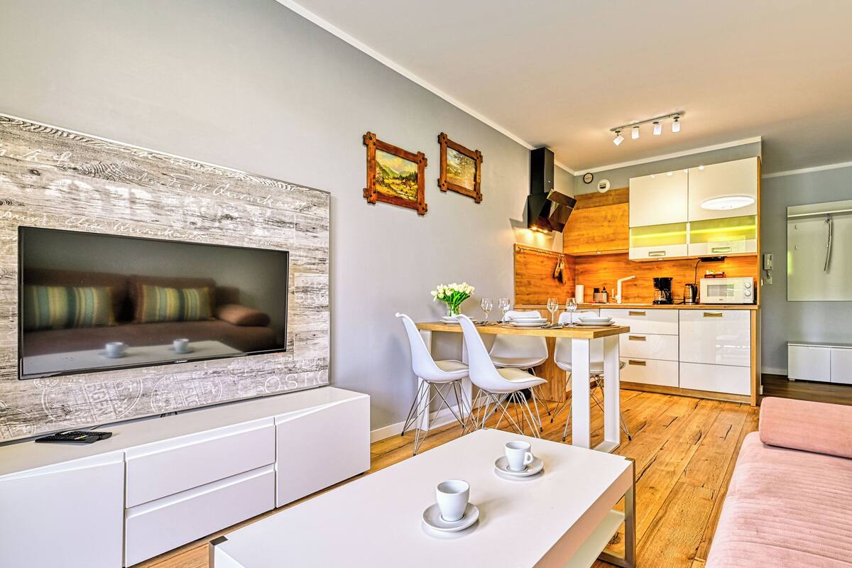 Ein modernes Wohnzimmer mit Flachbildfernseher, einem Essbereich mit Holztisch, weißen Stühlen und einem rosafarbenen Sofa.