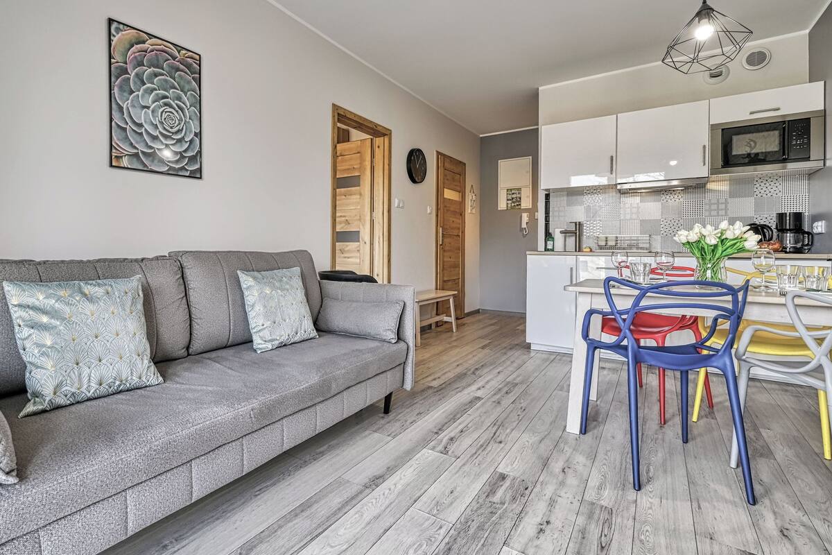 Ein modernes Wohnzimmer mit grauem Sofa, ein Essbereich mit bunten Stühlen und eine Küche mit weißen Schränken und einer Mikrowelle.