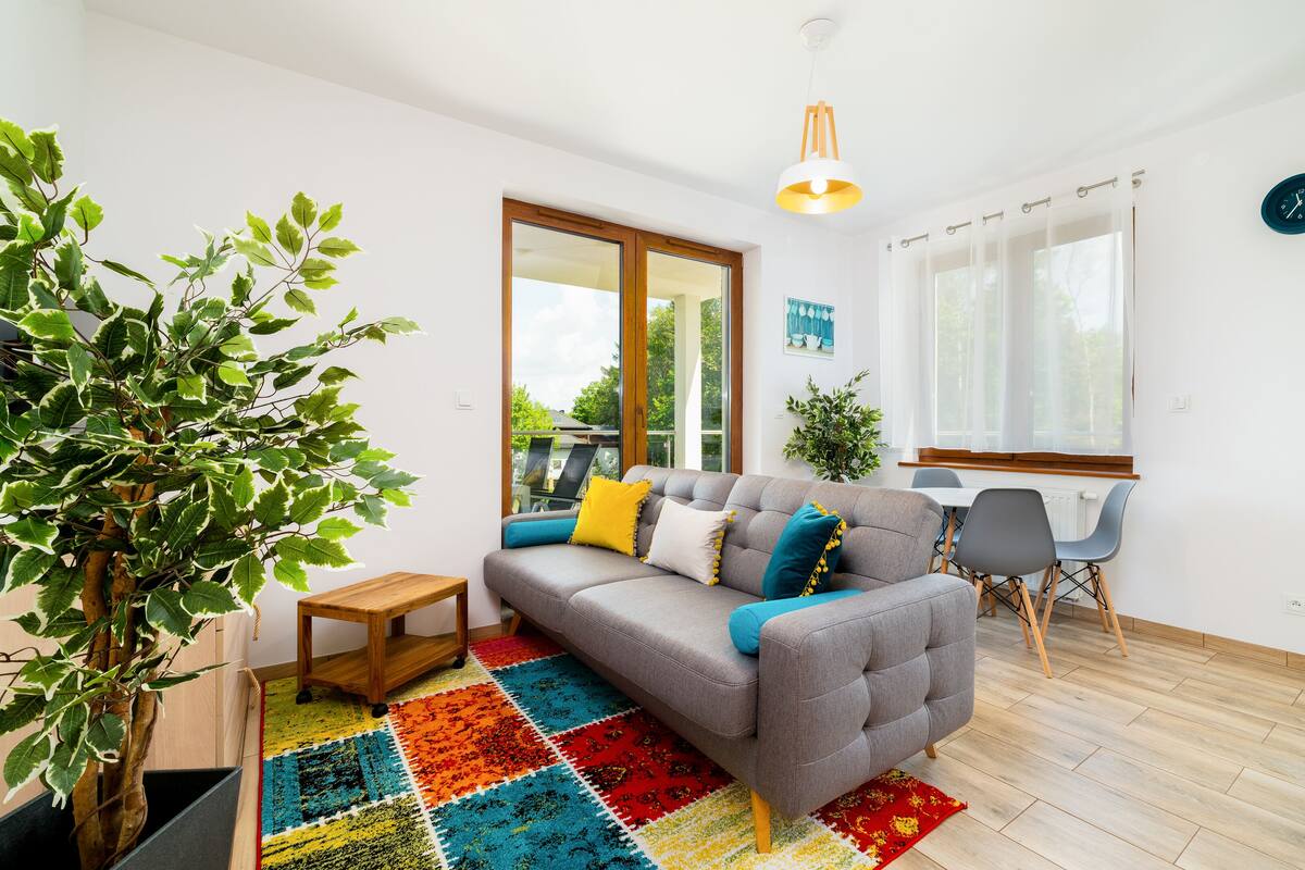 Ein modernes Wohnzimmer mit einem grauen Sofa, einem farbenfrohen Teppich, einem Holztisch und einer Topfpflanze.