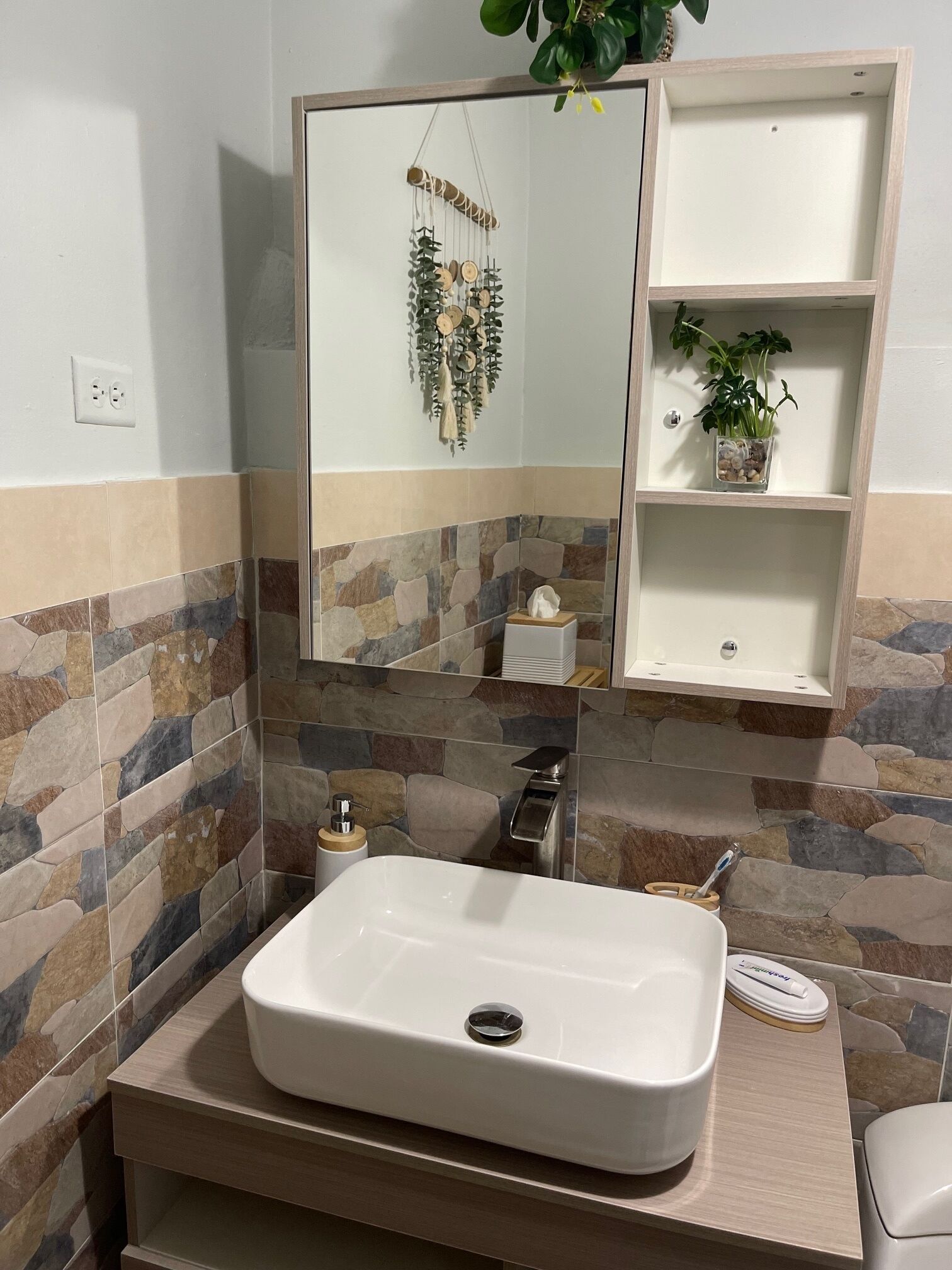 A bathroom with a stone-tiled wall, a white sink, a mirror, a shelf with a plant, and a toothbrush holder.