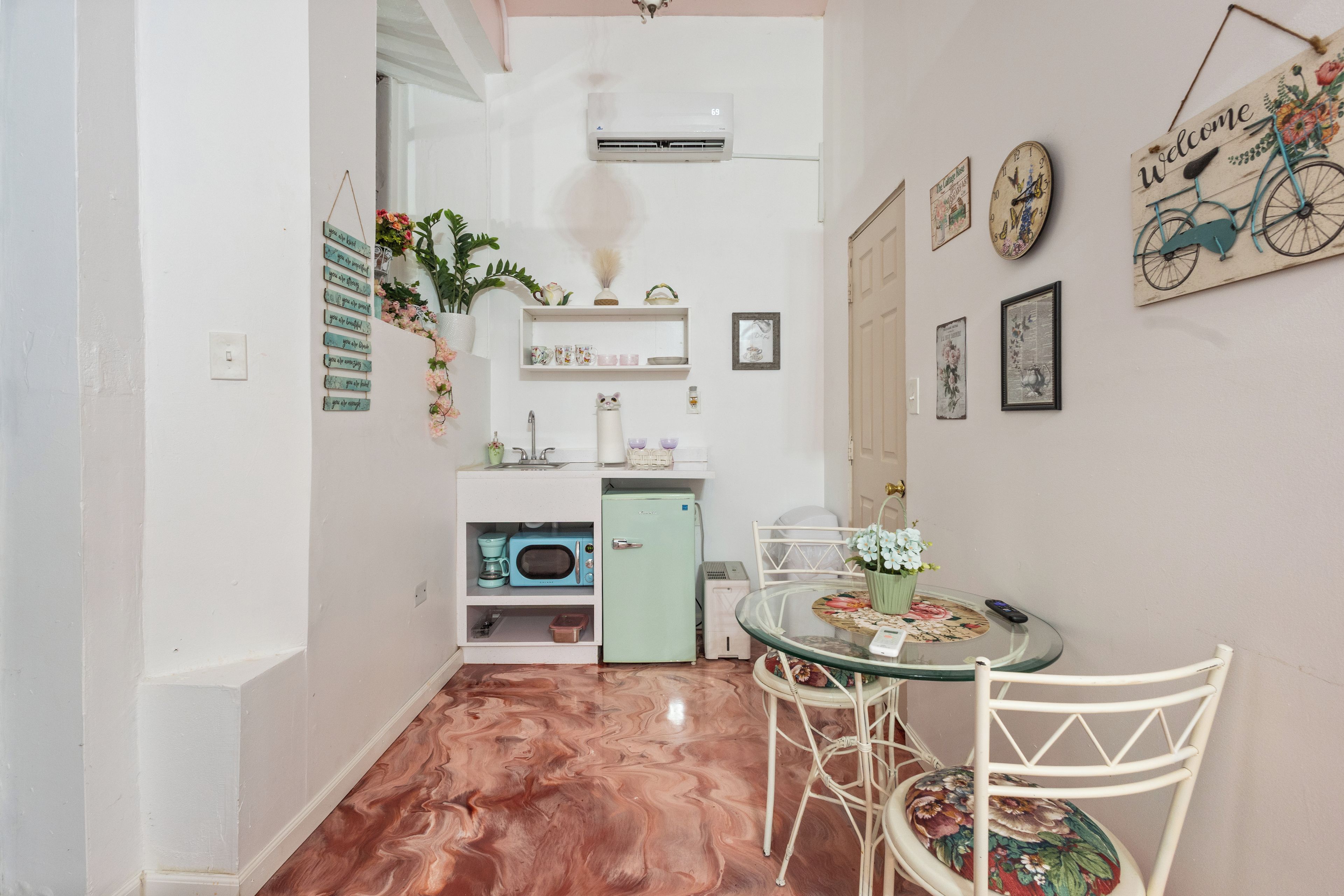 Uma cozinha compacta com micro-ondas, pequena geladeira e pia. Há uma área de refeições com mesa de vidro e cadeiras.