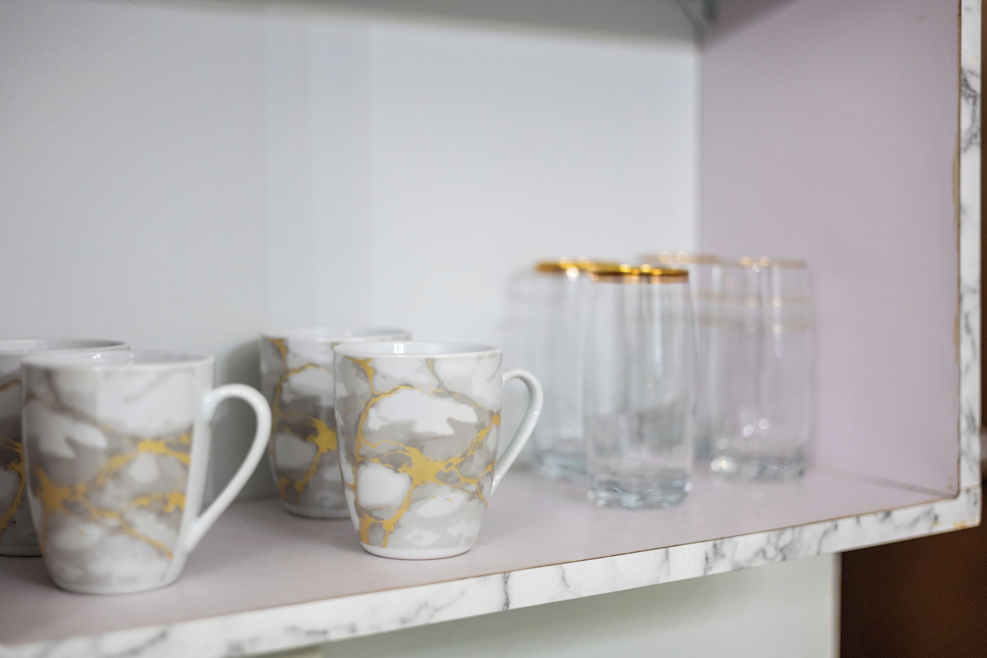 Una estantería con tazas de cerámica blanca con diseños dorados y recipientes de vidrio transparente.