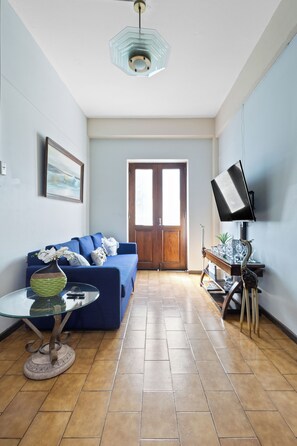 Ein Wohnzimmer mit einem blauen Sofa, einem Glastisch, einem Bild an der Wand und einem Fernseher auf einem Schrank.