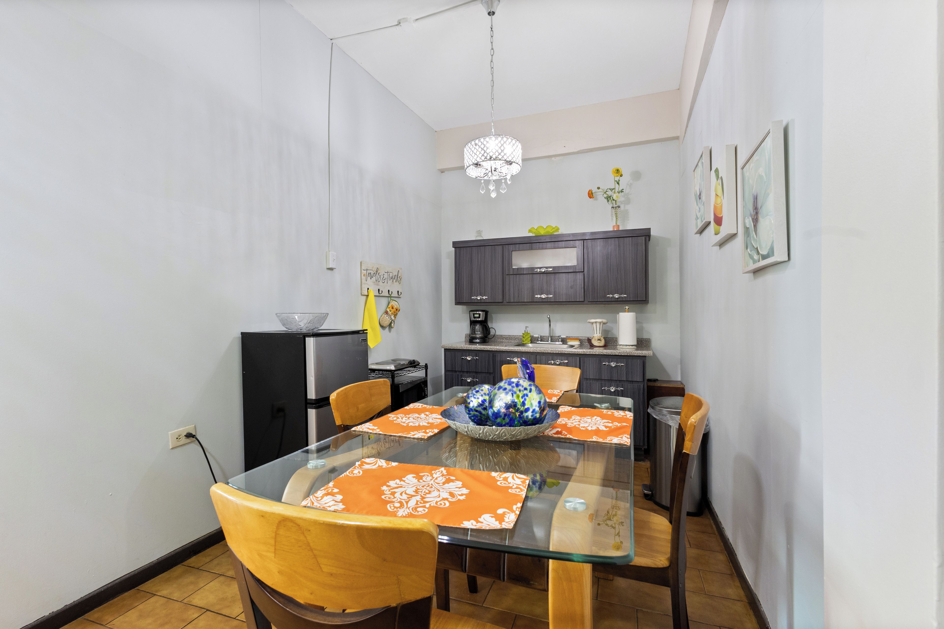 A compact dining area with a glass table, wooden chairs, a kitchen in the background, and a chandelier.