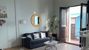 A modern living room with a dark grey sofa, a round coffee table, and a bed with a white duvet. A large mirror with a gold frame is on the wall, and there is a potted plant near the window. - Casa Of Essence (San Juan)