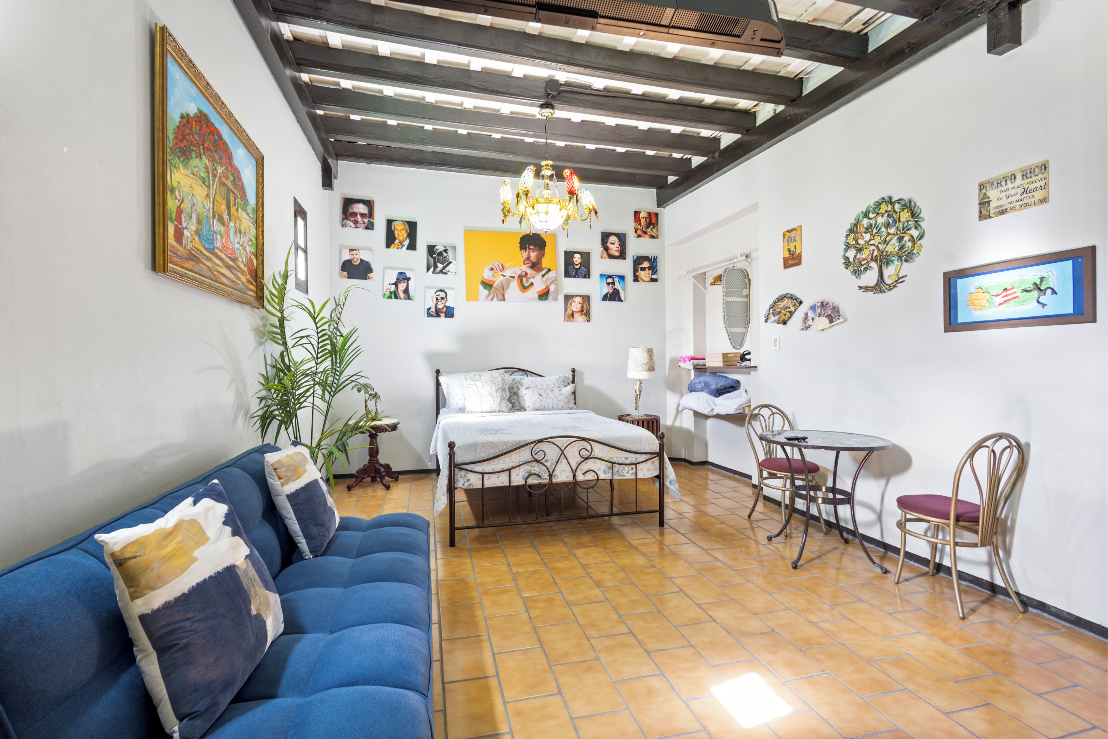 Un dormitorio con cama, sofá azul, mesa de comedor con sillas y diversas decoraciones en la pared, incluyendo fotografías y un cartel.