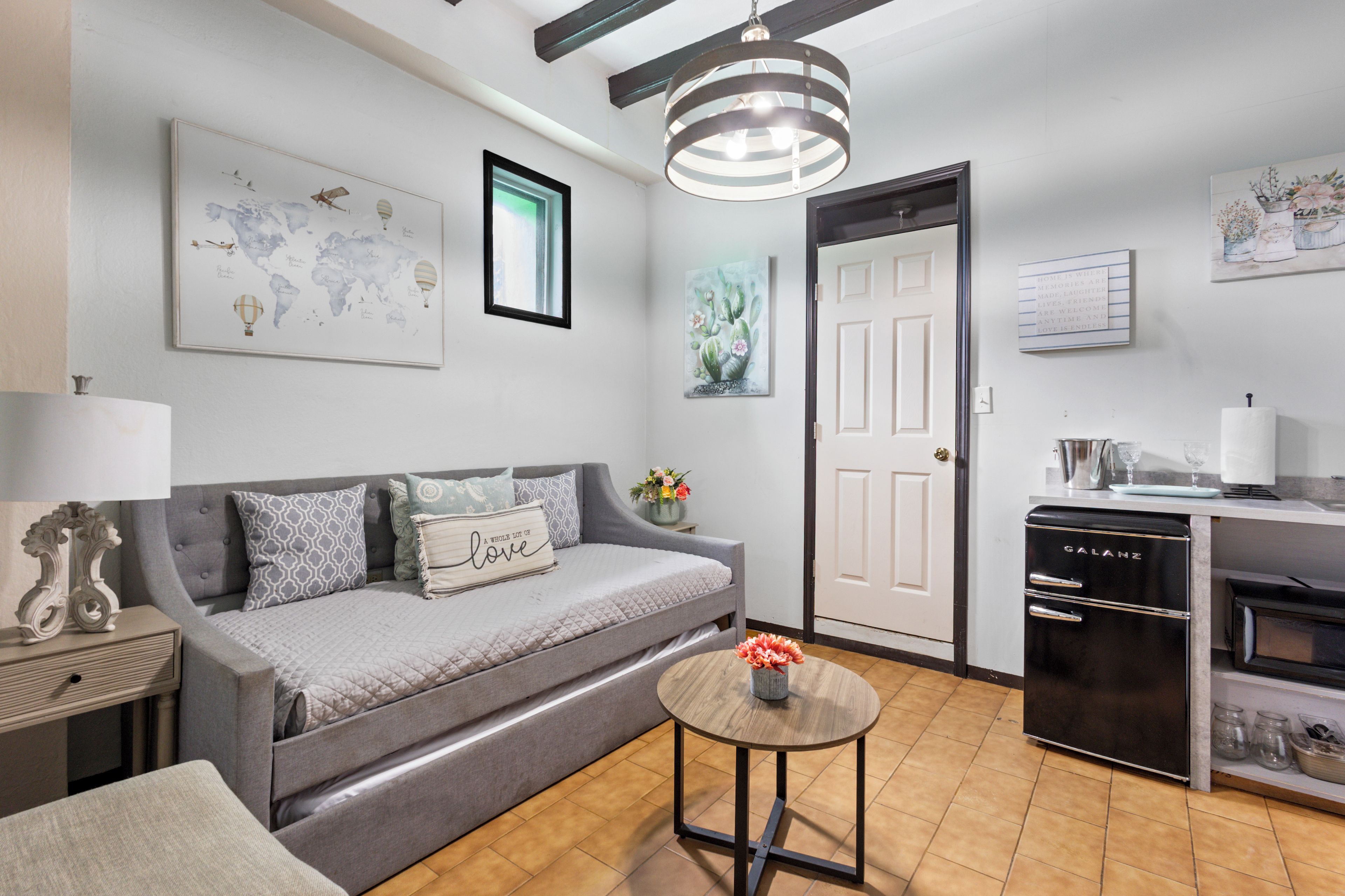A modern living room with a grey sofa, a small round table with a vase, and a kitchen area with a black refrigerator and a white door.