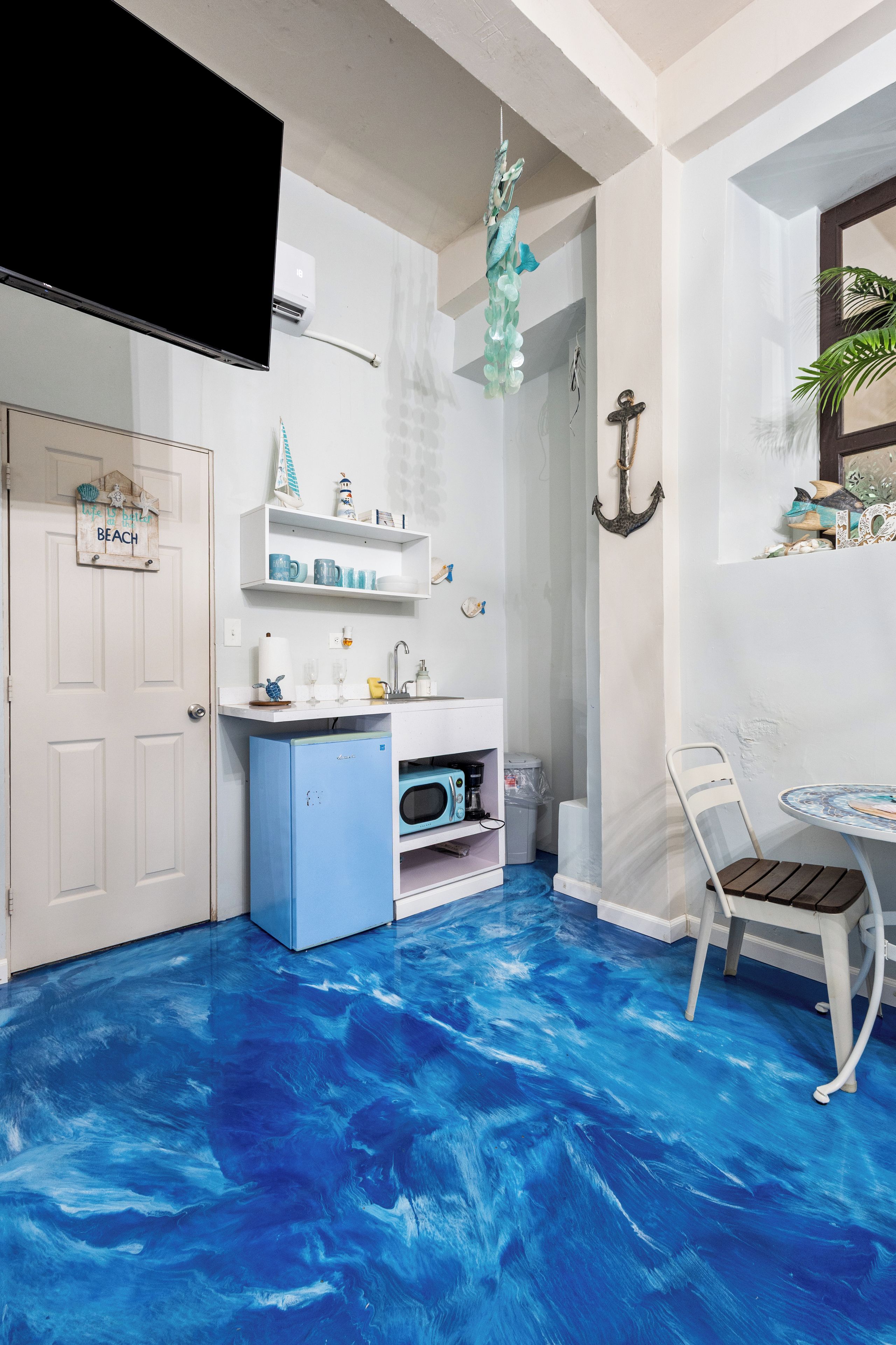 A room with a blue floor, white walls, a white door, a small kitchenette, a white chair, and a hanging light fixture.